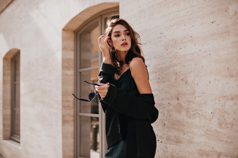 cute young girl with dark wavy hairstyle bright makeup silk dress black jacket holding sunglasses hands looking away against beige building wall 1